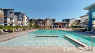 This contemporary residential community is defined by its striking multi-story buildings, finished in a bold palette of deep blue, warm gray, and natural wood-toned accents. The architecture features clean lines, pitched rooflines, and generous balconies that overlook the well-appointed outdoor amenity spaces below.

At the heart of the community lies a spacious resort-style swimming pool with crystal-clear turquoise water, complemented by a separate spa for relaxation. The expansive pool deck is outfitted with rows of lounge chairs and shade umbrellas, creating an inviting environment for sunbathing and outdoor leisure. A dedicated spa area provides an additional retreat near the pool.

Adjacent to the pool, a covered outdoor patio offers a shaded communal gathering space furnished with large dining tables, colorful seating, and an outdoor fireplace with a mounted screen — ideal for social gatherings in any season. The surrounding grounds feature thoughtfully designed landscaping with ornamental grasses, flowering shrubs, and mature trees, enclosed by a sleek black iron perimeter fence that adds both security and visual appeal.

Distant mountain views are visible from multiple vantage points throughout the property, enhancing the overall sense of openness. The combination of modern architecture, vibrant outdoor amenities, and carefully maintained green spaces creates a cohesive and resort-inspired living environment.