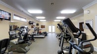 This fitness center offers a comprehensive workout environment thoughtfully designed for functionality and comfort. The space is finished with warm yellow walls, white crown molding, and a neutral carpeted floor, creating an inviting atmosphere that encourages regular use. Bright recessed panel lighting illuminates the entire room evenly, ensuring excellent visibility throughout all hours of the day. The facility is well-stocked with a range of cardio equipment, including treadmills, elliptical trainers, and a stair climber, all arranged along the perimeter to maximize open floor space in the center. Additional exercise accessories, such as a medicine ball, are available for varied workout routines. Wall-mounted flat-screen televisions are positioned for easy viewing during cardio sessions, adding an entertainment element to the exercise experience. Natural light filters in through several windows fitted with wooden blinds, complementing the overhead lighting. A set of double glass-paned doors at the far end of the room provides a welcoming entry point and contributes to the sense of openness. The single-floor layout ensures easy accessibility, making this fitness center a practical and well-appointed facility for a wide range of users.