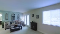 This single-floor space centers around a welcoming living room finished in soft, muted tones of sage green and light blue, creating a calm and airy atmosphere. The room is carpeted in a light neutral shade that complements the walls and contributes to an overall sense of warmth and comfort. Natural light flows generously into the space through a set of white-framed French doors, which likely lead to an outdoor area, as well as a large side window dressed with horizontal blinds. Together, these openings ensure the room remains bright throughout the day. The living area is thoughtfully laid out, with enough room to accommodate a full seating arrangement alongside a low-profile coffee table. A dark wood console cabinet adds a contrasting accent against the lighter walls and serves as a functional storage piece. Decorative framed artwork and wall-mounted pieces provide visual interest without overwhelming the space. The ceiling is smooth and painted white, enhancing the sense of openness. Overall, the space offers a comfortable and well-lit environment with a cohesive, understated design aesthetic.
