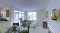 This single-floor space showcases a thoughtfully designed open-concept layout that seamlessly blends the living and dining areas into one welcoming environment. The interior is bathed in natural light, courtesy of large windows and a glass door that offer views of the surrounding outdoors, creating an airy and refreshing ambiance throughout.

The living area is generously proportioned, providing ample room for comfortable seating arrangements. A neutral color palette of soft whites and light grays enhances the sense of openness, while plush carpeting underfoot adds warmth and comfort. White upholstered ottomans serve as versatile accent pieces, complementing the relaxed yet polished atmosphere.

The dining area, positioned adjacent to the living space, features a sleek glass-top table paired with olive-green upholstered chairs, adding a touch of contemporary style and color contrast. A wood-toned sideboard along the wall provides functional storage while contributing to the room's warm, layered aesthetic.

Natural light streams in from multiple directions, highlighting the clean lines and uncluttered design of the space. The overall ambiance is calm, modern, and inviting — ideal for both everyday living and casual entertaining.