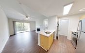 This single-floor unit is designed with a clean, modern aesthetic characterized by white walls, wood-look flooring throughout the main living areas, and an abundance of natural light. The open-concept layout connects the kitchen and living area seamlessly, creating a functional and airy feel.

The kitchen features a peninsula with a white quartz countertop and a stainless steel undermount sink equipped with a contemporary gooseneck faucet. Light wood cabinetry lines the walls, complemented by stainless steel appliances including a full-size refrigerator and range. The kitchen opens directly to the living area, which is anchored by a tile-surround fireplace and benefits from vaulted ceilings and a ceiling fan overhead. Sliding glass doors lead to a private balcony with lush outdoor views, enhancing the sense of connection to the surroundings.

The bedroom is carpeted for comfort and includes a ceiling fan, a generously sized window overlooking greenery, and ample wall space. The bathroom is finished in a crisp white palette, with a single-sink vanity topped with a white countertop, a large mirror, and a full tub-shower combination with a curved curtain rod. Wood-look flooring extends into the bathroom, tying the space together cohesively.