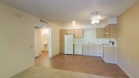 This single-floor space offers an open-concept layout that seamlessly connects the living area to the kitchen, creating a functional and cohesive environment. The living area features warm wood-style laminate flooring, transitioning to carpet near the entry zone, and is illuminated by a ceiling fan with an integrated light fixture, providing both airflow and ambient lighting.

The kitchen is positioned along the back wall and is equipped with light wood cabinetry offering ample upper and lower storage. White appliances include a full-size refrigerator, a dishwasher, an electric stovetop range, and a double basin sink with a window positioned above it. A ventilation hood is mounted above the range, and additional open shelving adds practical storage options.

The textured popcorn ceiling runs throughout the space, and a central HVAC vent ensures climate control. A doorway visible from the main living area leads to a bathroom, which features a wall-mounted sink and mirror, hinting at a compact but efficient layout. The overall color palette of warm beige and neutral whites gives the space a clean, modest aesthetic.