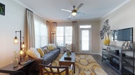 Located at 1680 Bluewater Way in Charleston, South Carolina, this single-floor residence offers a thoughtfully designed living experience with a warm and inviting atmosphere. The living area serves as the heart of the home, showcasing hardwood-style flooring, crown molding, and neutral wall tones that create a clean, versatile backdrop. Large windows dressed with elegant draping panels allow an abundance of natural light to fill the room, enhancing its open and airy feel. A ceiling fan with a built-in light fixture provides both comfort and functionality overhead. The space opens directly to an exterior door, suggesting access to a private outdoor area. The living room is generously proportioned, comfortably accommodating a full seating arrangement centered around a rustic coffee table. Warm yellow and grey accent tones carry throughout the decor, lending the space a cohesive and stylish character. The overall ambiance of this Charleston residence is bright, modern, and livable, blending practical design with welcoming details that make it an ideal home in one of South Carolina's most sought-after communities.