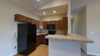 Located at 2066 Home Road in Grove City, Ohio, this single-story space offers a functional and inviting layout. The kitchen serves as a central highlight, showcasing rich medium-toned wood cabinetry that extends to the upper walls, providing generous storage throughout. Light granite-style countertops complement the warm cabinetry tones, while a peninsula counter extends outward, creating an ideal space for casual dining or food preparation. Black appliances, including a refrigerator, range, and over-the-range microwave, lend a cohesive, modern contrast to the warm wood tones. Recessed lighting is installed in a slightly raised ceiling section above the kitchen, ensuring the workspace is well-lit. Light hardwood-style flooring runs throughout the area, adding warmth and continuity. A partial wall and open sightlines connect the kitchen to adjacent living areas, contributing to an open, flowing feel. The neutral wall tones throughout keep the space feeling bright and adaptable, offering a versatile backdrop for a variety of interior styles. Overall, the space presents a clean, practical design well-suited for everyday living.