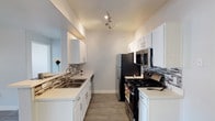 This single-floor space showcases a thoughtfully designed galley kitchen that serves as its centerpiece. The kitchen features crisp white shaker-style cabinetry lining both sides, providing ample storage, complemented by light stone countertops that offer generous prep space. A decorative mosaic tile backsplash in neutral tones adds a refined visual detail along the walls, tying together the clean, contemporary aesthetic.

The kitchen is equipped with a full suite of stainless steel appliances, including a gas range, over-the-range microwave, refrigerator, and dishwasher, making it both functional and stylish. A double-basin stainless steel sink with a modern pull-down faucet is conveniently positioned on one side, with natural light filtering in from a nearby window.

Underfoot, wood-look flooring runs throughout, lending warmth and continuity to the space. Track lighting mounted on the ceiling provides focused illumination across the kitchen workspace, ensuring both practicality and ambiance. The soft, neutral gray walls keep the overall palette light and airy, creating a welcoming environment. The layout is efficient and well-organized, maximizing every inch of available space while maintaining an open, uncluttered feel throughout.