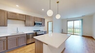 This well-appointed 2-bedroom, 2-bathroom home in Middleton, Wisconsin offers a clean, modern aesthetic across a single level. The open-concept kitchen and living area serves as the heart of the home, where warm hardwood-style flooring flows seamlessly throughout. The kitchen is outfitted with taupe shaker-style cabinetry, crisp white quartz countertops, a central island with pendant lighting, and a full suite of stainless steel appliances including a refrigerator, range, microwave, and dishwasher. The living area extends to a private balcony through sliding glass doors, offering pleasant outdoor views of the surrounding neighborhood. Both bedrooms feature plush carpeting, ceiling fans, and windows that invite natural light. The primary bedroom is generously sized and connects to a dedicated bathroom with wood-toned vanity cabinetry and light flooring. The second bathroom mirrors the modern design language with a clean white vanity, large mirror, and a full soaking tub. Two closets provide practical storage throughout the home. Warm wood trim accents and neutral walls create a cohesive, inviting atmosphere that carries from room to room.