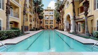 This single-floor residence is situated within a Mediterranean-inspired multi-story complex, characterized by warm yellow stucco exteriors, arched colonnades, and lush palm tree-lined courtyard corridors. The community's centerpiece is a generously sized outdoor swimming pool, framed by lounge seating and mature palms, evoking a resort-like atmosphere under open skies.

Inside, the interior design makes a confident statement through a carefully curated blend of materials and finishes. The dining area serves as a focal point, anchored by a substantial round green marble table with a pedestal base, surrounded by slender rattan-style chairs atop a woven area rug. The polished concrete flooring adds an industrial edge that contrasts elegantly with the warmth of the furnishings. Neutral white walls provide a refined backdrop for framed artwork, including a large-scale abstract piece in soft earth tones that draws the eye immediately upon entering.

A distinctive weathered-metal framed glass door connects the dining space to an adjoining room, adding an eclectic, artisanal touch to the overall aesthetic. A sleek barn door on a matte black rail system offers flexible room division while reinforcing the space's modern sensibility. Recessed lighting throughout ensures even, functional illumination, while the clean lines and understated palette create a calm, sophisticated ambiance throughout.