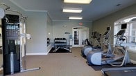 This fitness center offers a clean, functional environment designed to support a well-rounded workout routine. The space is finished with soft sage-green walls, white crown molding, and neutral carpeted flooring, creating a calm and welcoming atmosphere. Overhead, flush-mounted fluorescent light panels provide consistent, even illumination throughout the room. A set of elegant arched French doors along the far wall allows natural light to filter in while offering a view to the exterior, adding an airy quality to the interior. The cardio section is well-appointed with multiple Precor elliptical trainers and additional upright cardio machines, all positioned for ease of use and spacing. A dedicated free weights area features a tiered dumbbell rack stocked with a full range of weights, accompanied by an adjustable workout bench and a rubber floor mat for safety and stability. A water cooler is conveniently located near the entrance for easy access during workouts. The overall layout is thoughtfully organized to allow comfortable movement between equipment zones. With its blend of practical amenities, natural light, and tidy presentation, this fitness center provides a well-maintained and motivating space for residents or members to pursue their fitness goals.