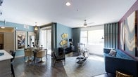 This single-floor space showcases a sophisticated, open-concept layout that seamlessly blends the living and dining areas into one cohesive environment. The interior is defined by striking teal accent walls that create a rich, dramatic backdrop, complemented by dark hardwood flooring that runs throughout, grounding the space with warmth and depth.

The living area benefits from large windows dressed with floor-length gray curtains, allowing generous natural light to pour in while offering views of an outdoor balcony. A ceiling fan with a decorative fixture enhances both function and style overhead. The space is furnished with a mix of bold, eclectic pieces — including a striped accent chair, a velvet blue sofa, and a glass-and-gold coffee table — that reflect a curated, contemporary aesthetic.

The dining area, partially separated by a column detail, features an ornate chandelier and a dark, refined dining set with metallic-accented chairs. A large decorative mirror adds depth to the space, while gold sculptural wall art provides eye-catching focal points throughout. The overall ambiance is elegant, bold, and carefully designed with a strong sense of style and personality.