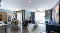 This single-floor space showcases a sophisticated, open-concept layout that seamlessly blends the living and dining areas into one cohesive environment. The interior is defined by striking teal accent walls that create a rich, dramatic backdrop, complemented by dark hardwood flooring that runs throughout, grounding the space with warmth and depth.

The living area benefits from large windows dressed with floor-length gray curtains, allowing generous natural light to pour in while offering views of an outdoor balcony. A ceiling fan with a decorative fixture enhances both function and style overhead. The space is furnished with a mix of bold, eclectic pieces — including a striped accent chair, a velvet blue sofa, and a glass-and-gold coffee table — that reflect a curated, contemporary aesthetic.

The dining area, partially separated by a column detail, features an ornate chandelier and a dark, refined dining set with metallic-accented chairs. A large decorative mirror adds depth to the space, while gold sculptural wall art provides eye-catching focal points throughout. The overall ambiance is elegant, bold, and carefully designed with a strong sense of style and personality.