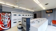 This shared laundry facility is designed for efficiency and ease of use, offering a well-organized layout that accommodates a high volume of laundry needs. The room features a row of stacked commercial-grade dryers along the back wall, providing ample drying capacity. Opposite, a lineup of top-load washing machines offers convenient access. A centrally positioned coin change machine ensures users can readily obtain the currency needed to operate the machines. The space is finished with neutral grey walls, creating a clean and functional atmosphere. Overhead, fluorescent lighting panels and ceiling fans provide bright, even illumination throughout the room while maintaining comfortable air circulation. The light-toned flooring complements the white appliances, contributing to an overall sense of cleanliness and order. A single entry door provides straightforward access to the facility. Smoke detectors are visibly installed on the ceiling, reflecting attention to safety. The space is practical and purposefully laid out, making it a convenient and fully equipped laundry resource.