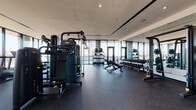 This contemporary fitness center is designed to deliver a premium workout experience within a sleek, modern setting. The space is defined by expansive floor-to-ceiling glass curtain walls that line the perimeter, flooding the interior with natural light and offering impressive panoramic views of the surrounding city skyline — making even routine workouts visually engaging.

The flooring throughout is finished in durable rubber matting, providing both safety and comfort underfoot. The fitness floor is comprehensively outfitted with a wide array of professional-grade equipment. Cardio options include multiple treadmills and an elliptical trainer, all positioned directly facing the large windows to take full advantage of the cityscape views. The strength training section features a multi-station cable machine, a Smith machine with weight plate storage, a full rack of dumbbells ranging across various weights, adjustable workout benches, and a selection of medicine balls and kettlebells.

Full-length mirrors are mounted along one wall, enhancing the sense of space and providing functional use during workouts. The entry area transitions through an open corridor featuring exposed concrete columns and polished terrazzo-style flooring, giving the space an industrial-modern architectural character. Recessed ceiling lighting throughout ensures the facility remains bright and well-illuminated at all times.