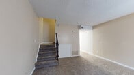 Located at 7000 Barrington Dr in Charlotte, NC, this two-floor home offers a straightforward and functional layout across two connected levels. The main floor greets visitors with a carpeted living area bathed in natural light from a large window, complemented by a six-panel front entry door and a staircase with a black metal railing leading to the upper level. The textured ceilings throughout lend a classic residential character to the interior. The kitchen features white cabinetry, light countertops, a double stainless steel sink, and a black dishwasher, with hardwood-style flooring adding warmth underfoot. Upstairs, the second floor continues with wall-to-wall carpeting and neutral beige-toned walls, providing a calm and comfortable atmosphere. The bedroom is a generously sized room with a closet and ample space for furnishings. The bathroom is finished in crisp white tile wainscoting, diamond-pattern floor tile, a white vanity with under-cabinet storage, a mirrored medicine cabinet, and clean fixtures throughout. The upper level also features a spacious open area with HVAC vents, offering flexible use of space. Overall, the home presents a clean, move-in ready interior with consistent neutral tones and practical finishes throughout.