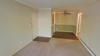 This single-floor residence offers a thoughtfully laid-out living space that combines comfort and classic design details. The entryway opens directly into a carpeted living area, providing a warm and welcoming first impression. Crown molding trims the ceiling throughout, adding a refined architectural touch to the interior.

Transitioning further into the home, the flooring shifts to rich, dark hardwood-style planks that extend through the dining area and connecting spaces, creating a clear visual distinction between zones. The dining area features a ceiling fan, offering both functional air circulation and a casual, homey ambiance. The walls are finished in a two-tone style, with a neutral upper half and a muted sage green lower half divided by a chair rail, lending the space a traditional yet cozy character.

A glimpse into the adjacent kitchen reveals warm cabinetry and a tiled backsplash, hinting at a functional and well-appointed cooking space. The open sightlines between the living, dining, and kitchen areas contribute to a sense of spaciousness and ease of movement throughout the home. Overall, the space is characterized by its classic detailing, cohesive color palette, and practical layout suited for everyday living.