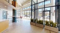 Located at 337 Seventh St W in Saint Paul, Minnesota, this sophisticated two-story commercial space offers a striking blend of industrial and contemporary design across a generous footprint. The main level commands attention with soaring double-height ceilings, expansive floor-to-ceiling glazing that floods the interior with natural light, and polished concrete floors that lend an urban, refined character. A grand lobby greets visitors with sculptural wire pendant lights, dual elevators framed by textured concrete walls, and a curated gallery-style art wall — setting a distinctly creative tone from the moment of entry. Sliding glass partitions divide flexible lounge and meeting areas, furnished with modern seating arrangements that invite both collaboration and casual interaction. A standout teal-glazed tile column anchors a central gathering spot beside a light wood kitchenette, while a bold, colorful mural wraps a feature wall, adding vibrant artistic energy to the space. The staircase, accented with warm wood slat detailing and cable railings, leads to the upper level, where light hardwood flooring, exposed white ductwork, and an open-plan layout create an airy, loft-like atmosphere. A built-in bar counter with leather stools and multiple lounge seating clusters make the upper level ideal for both focused work and social engagement, with views over the surrounding neighborhood completing the experience.