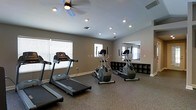 This single-floor fitness room offers a dedicated space for cardiovascular exercise and general wellness. The room is outfitted with multiple treadmills and elliptical cross-trainers, providing a variety of cardio workout options. A large wall-mounted mirror reflects the space, enhancing the sense of openness while allowing users to monitor their form during exercise. Along the far wall, a low-profile shelving unit offers convenient storage for personal items and fitness accessories. The flooring features durable, rubberized material well-suited for a gym environment, providing both cushioning and easy maintenance. Natural light filters in through multiple windows fitted with blinds, complementing the recessed ceiling lights and a ceiling fan that work together to keep the space well-lit and ventilated. The neutral gray walls and clean white trim create a calm, focused atmosphere conducive to exercise. The overall layout is thoughtfully arranged to allow comfortable movement between equipment, making this fitness room a practical and inviting facility for daily workouts.