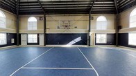Located at 100 Delaney Dr in Jacksonville, NC, this single-floor indoor sports facility is purpose-built for athletic activity and recreation. The centerpiece of the space is a full-size basketball court finished in a deep blue sport tile flooring, complete with clearly marked court lines and a regulation-height glass backboard with rim and net mounted to the far wall. The soaring ceiling height, supported by an exposed steel beam framework, provides ample clearance for play and contributes to the expansive, open feel of the interior. Natural light pours in generously through large arched windows positioned high on the walls on both sides of the court, creating a bright and welcoming atmosphere throughout the day. Industrial ventilation fans are mounted near the upper walls to ensure proper airflow. The lower portion of the walls features a navy blue wainscoting treatment that contrasts with the cream-toned upper walls, giving the space a clean and polished aesthetic. Entry doors are accessible from multiple points along the perimeter. Overall, the facility combines functional athletic infrastructure with a well-maintained, professional environment suited for basketball, recreational sports, or multi-purpose fitness use.