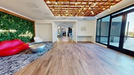 Located at 480 Morrissey Blvd in Boston, MA, this single-floor commercial space offers a generously sized, thoughtfully designed environment that blends creative aesthetics with functional versatility. The entry area immediately captivates with warm hardwood-style flooring, a striking wood-lattice ceiling adorned with Edison bulb string lights, and a living green wall framed in wood — accented by a bold red lip-shaped sofa, a metallic coffee table, and a neon sign reading "your own reality," setting an energetic and artistic tone throughout.

The open-plan layout transitions seamlessly into a spacious communal area featuring polished concrete floors, exposed industrial ceilings with geometric pendant lighting, and an integrated kitchenette with a large island. A substantial conference table with designer seating and a bold black-and-white patterned rug anchors a dedicated meeting zone. Nearby, a lounge area with modular sectional seating and a mounted display screen provides a relaxed gathering space.

Adding a playful dimension, an arcade gaming corner with retro machines and vibrant pink booth seating brings a fun, social character to the space. Private enclosed meeting pods with glass accents and soft ambient lighting offer quieter work options. Large windows throughout invite natural light and views of the surrounding Boston neighborhood, completing this dynamic and inspiring environment.