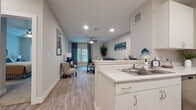 This single-floor residence offers a thoughtfully designed open-concept layout that seamlessly connects the kitchen, dining, and living areas. The kitchen is equipped with stainless steel appliances — including a refrigerator, range, and over-the-range microwave — complemented by crisp white cabinetry, light quartz countertops, and wood-look luxury vinyl plank flooring. A breakfast bar with seating provides a convenient casual dining option and serves as a natural transition into the living space. The living area is bright and welcoming, featuring a ceiling fan, recessed lighting, and large windows dressed with decorative drapes that invite natural light while offering privacy. The bedroom is finished with plush carpeting for added comfort and includes a dedicated walk-in closet with built-in shelving and hanging storage. The full bathroom is well-appointed with a tub-shower combination featuring tile surround, a white vanity with storage, and grab bars for added accessibility. Throughout the residence, neutral tones, clean lines, and consistent finishes create a cohesive and modern aesthetic that is both functional and inviting.