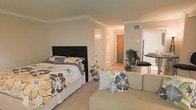This single-floor studio efficiently combines living and sleeping areas within one open space. The main room features carpeted flooring and is finished with elegant crown molding that adds a refined touch to the neutral beige walls. A dedicated sleeping area accommodates a full-sized bed with a bold black headboard, complemented by decorative bedding in yellow, gray, and white floral patterns. Adjacent to the sleeping area, a compact seating area with a cushioned sofa and coordinating accent pillows in matching tones creates a defined lounging space.

A hallway visible from the main room leads to additional areas of the unit, including what appears to be a closet and an entryway with a wood-toned door. Hardwood-style flooring runs through the hallway and extends into a kitchen nook area, where a tall bistro-style table and chairs are partially visible alongside what appears to be a refrigerator. A potted indoor plant near the transition between spaces adds a natural element to the interior. Recessed HVAC vents and overhead lighting contribute to a comfortable, functional environment throughout.