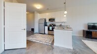 Located on Bridgeport Way in Suffolk, Virginia, this single-floor residence offers a thoughtfully designed layout that balances comfort and modern style. The open-concept kitchen is equipped with stainless steel appliances, including a refrigerator, range, and microwave, complemented by crisp white cabinetry, granite countertops, and a peninsula with pendant lighting overhead. Polished concrete flooring flows through the kitchen and into the living area, lending a contemporary industrial character to the space. The living room is generously sized, bathed in natural light from multiple large windows fitted with white blinds, and offers a welcoming atmosphere with neutral-toned walls that enhance the airy feel. The bedroom is carpeted for warmth and comfort, featuring a large window with outdoor views that fill the room with soft, natural light. A well-sized closet provides ample storage. The bathroom showcases granite countertops atop white shaker-style cabinetry, a large vanity mirror, and hardwood-style flooring, combining clean aesthetics with practical functionality. Throughout the residence, the neutral color palette, modern finishes, and abundant natural light create a cohesive and inviting living environment suited for everyday comfort.
