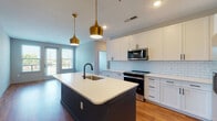 This contemporary single-floor residence showcases a thoughtfully designed open-concept layout that seamlessly blends style and functionality. The kitchen serves as the centerpiece of the home, featuring crisp white shaker cabinetry, a classic white subway tile backsplash, and stainless steel appliances including a range and over-the-range microwave. A striking two-tone island with a dark teal base and white quartz countertop anchors the space, complemented by a matte black faucet and sink. Paired gold pendant lights hang above, adding a warm, sophisticated touch. Rich hardwood flooring flows throughout the main living areas, enhancing the home's warm ambiance. Natural light pours in through large windows fitted with blinds, offering views of the surrounding area. The bedrooms are well-proportioned, each equipped with ceiling fans and large windows. The primary bathroom is finished with large-format tile flooring, white shaker vanity cabinetry, a framed mirror, and matte black fixtures, with direct access to a generously sized walk-in closet outfitted with wire shelving systems. A second bathroom features a subway-tiled tub-shower combo with matching finishes. Additional walk-in closet space adds practical storage throughout this well-appointed home.