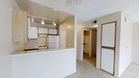 This single-floor residence is designed with an open-concept layout that connects the kitchen and living areas seamlessly. The kitchen is equipped with white appliances, including a refrigerator and an electric range with overhead microwave cabinetry. A peninsula-style breakfast bar with a white countertop creates a natural division between the kitchen and the adjacent living space, offering both additional workspace and a casual dining option. Track lighting is installed above the kitchen, providing focused, adjustable illumination across the cooking and prep areas. The ceiling features a textured finish throughout, and a ceiling fan is positioned in the main living area to promote air circulation. The living area is finished with wall-to-wall carpet, adding warmth and softness underfoot. A hallway visible from the main space leads to a bathroom, which appears to include a shower and a walk-in or reach-in closet with hanging storage. Neutral beige and white tones dominate the interior palette, giving the space a clean, cohesive look. HVAC vents are integrated into the ceiling, ensuring climate control throughout. The overall layout is functional and efficiently organized for everyday living.
