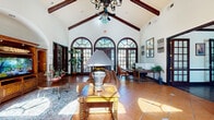 This single-story space presents a compelling blend of Spanish-Colonial architectural character and functional design across a well-proportioned floor plan. The centerpiece is a dramatic main living area distinguished by soaring vaulted ceilings accented with dark-stained exposed wood beams and an ornate chandelier fan. A sweeping wall of arched, multi-pane French doors floods the space with abundant natural light while offering views of lush outdoor greenery, reinforcing an open, airy ambiance. Warm terracotta tile flooring grounds the room and complements the rich, dark wood trim found throughout.

The kitchen features dark espresso cabinetry, granite countertops, a wraparound breakfast bar with woven bar seating, stainless steel appliances, and a tile backsplash, creating a stylish and functional culinary space.

The dedicated office is accessed through French doors and offers hardwood-style flooring, large windows with wood blinds allowing ample natural light, and sufficient workspace for productivity in a calm setting.

A fitness room provides space for cardiovascular and strength equipment, lined with multiple windows and ceiling fans for ventilation. Throughout the space, dark wood trim, arched entryways, and thoughtful transitions between rooms create a cohesive and inviting atmosphere.