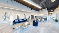 Located at 44 High St in downtown Nashua, New Hampshire, this contemporary commercial space offers a thoughtfully designed, single-floor open-plan layout. The interior is defined by soaring ceilings with exposed dark wood beams, visible ductwork, and sleek linear pendant lighting that lends an industrial-chic aesthetic balanced by refined modern furnishings.

Polished concrete floors run throughout, reflecting natural light that pours in from an expansive wall of floor-to-ceiling windows along the street-facing façade. These oversized windows provide panoramic views of the surrounding downtown streetscape while flooding the interior with daylight.

The main gathering area is furnished with bold navy blue velvet sofas and curved accent chairs, anchored by sculptural metallic coffee tables atop a layered area rug. Adjacent to the lounge is a communal dining area with a warm wood table surrounded by contemporary white chairs, illuminated by large square pendant fixtures. A well-appointed kitchenette with blue cabinetry and open shelving adds functional elegance.

Glass-walled private rooms provide flexibility for focused work or meetings. Along a secondary corridor, bench seating lines the window wall, creating a calm, light-filled passage. Curated abstract artwork adorns the walls throughout, tying together the blue and neutral color palette. The overall ambiance is sophisticated, vibrant, and purposefully designed for a dynamic professional environment.