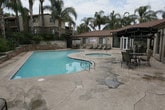 This outdoor amenity area is designed to deliver a resort-like experience within a gated residential community. The centerpiece is a generously sized swimming pool with a freeform shape, featuring vivid turquoise water and step-in entry with stainless steel handrails. Adjacent to the pool sits a circular spa, offering a dedicated space for relaxation. The surrounding concrete pool deck provides ample space for lounging, with sling-style chaise lounge chairs positioned to take advantage of the open sky and sunshine.

The landscaping plays a prominent role in the ambiance of this space. Tall, mature palm trees frame the area, complemented by layered plantings of tropical shrubs and flowering plants set within raised stone-faced retaining walls. The warm-toned stucco buildings surrounding the courtyard feature balconies and tile-accented rooflines, reinforcing a Mediterranean-influenced architectural style.

An outdoor shower station is integrated into the space near the pool entrance, finished entirely in charcoal grey mosaic tile with a curved interior wall and a wall-mounted showerhead. A gated entry with ornamental ironwork provides secure access, completing this well-appointed and thoughtfully designed outdoor recreational area.