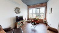 Located at 509 Market St in the heart of downtown Shreveport, Louisiana, this single-floor urban space showcases a compelling blend of industrial heritage and contemporary design. The space is defined by its prominent exposed brick walls, which appear throughout multiple rooms, including the living area and one of the bathrooms, lending a raw, historic texture that contrasts beautifully with crisp white walls and dark-painted ceilings.

The living area is a standout, featuring soaring ceilings, rich hardwood-style flooring, and oversized windows that frame sweeping views of the downtown Shreveport skyline. Natural light floods the room, highlighting the eclectic character of the space.

The kitchen is well-appointed with two-tone cabinetry — dark lower cabinets paired with white uppers — complemented by light countertops, stainless steel appliances, and gold-toned fixtures that add a warm, modern accent. The high ceilings continue here, reinforcing the open, airy feel.

Two bedrooms offer distinct personalities: one features exposed brick and large windows with street views, while the second is a quieter, more intimate room with tall ceilings and a built-in wardrobe. Two bathrooms serve the space, one with a glass-enclosed shower alongside an exposed brick accent wall, and another featuring a dark-framed vanity with gold fixtures.