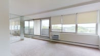 This single-floor space is characterized by its bright, open layout with an abundance of natural light streaming through large windows and sliding glass doors that line an entire wall. The sliding glass doors open to a balcony with a decorative lattice railing, offering views of lush, tree-lined surroundings. Roller shades in a neutral beige tone are installed across the windows, providing flexible light control and privacy. The flooring throughout is covered in wall-to-wall carpet in a soft, neutral tone that complements the clean, light-colored walls and ceiling. A window-mounted air conditioning unit is installed beneath one of the windows, ensuring climate comfort. The wall of glass creates a seamless connection between the interior and the outdoor environment, making the space feel airy and expansive. A mirrored wall panel is also present, further enhancing the sense of openness and reflecting the natural light throughout the room. Baseboard heating runs along the perimeter, adding to the practical functionality of the space. Overall, the room presents a clean, minimalist aesthetic with an emphasis on natural light and outdoor connectivity.