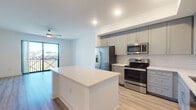 This well-appointed single-floor residence showcases a thoughtfully designed open-concept layout with consistent light wood-tone flooring flowing throughout. The kitchen serves as a central highlight, featuring gray shaker-style cabinetry extending to the upper walls, a classic white subway tile backsplash, stainless steel appliances including a French door refrigerator with ice maker, a range, microwave, and dishwasher, and a generous white quartz island that provides additional prep and seating space. Recessed lighting illuminates the kitchen area, while ceiling fans in both the kitchen-adjacent living zone and bedrooms enhance comfort and airflow.

The living area opens directly to a private balcony through large sliding glass doors, flooding the space with natural light and offering outdoor views. Additional bedrooms are bright and airy, each featuring light-filled windows with blinds and ceiling fans. The bathroom is finished with a walk-in glass-enclosed shower with white tile surround, gray shaker vanity with quartz countertop, a warm-toned framed mirror, and light wood-look flooring. Crisp white walls are consistent throughout, lending a clean, contemporary aesthetic to the entire space.