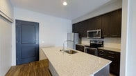 Located at 3443 Park Street in Grove City, Ohio, this single-floor residence offers a clean, modern aesthetic with thoughtfully selected finishes throughout. The kitchen serves as a central highlight of the space, showcasing a large island with light granite-style countertops and an undermount stainless steel sink with a sleek arc faucet — ideal for both meal preparation and casual dining. Rich dark cabinetry lines the walls, providing ample storage while contributing to the kitchen's sophisticated, contemporary look. Stainless steel appliances, including a refrigerator, electric range, and over-the-range microwave, complete the well-equipped cooking area. The flooring features warm-toned hardwood-style planks that add character and continuity throughout the space. Crisp white walls and recessed lighting create a bright, open atmosphere that enhances the sense of space. A bold dark entry door adds a striking design contrast at the entryway, framing the transition into the living area. Overall, the space combines functional design with modern finishes, making it a practical and visually appealing residence in Grove City.