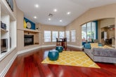 This single-floor space makes a striking first impression with its bold design choices and open, flowing layout. Rich, dark hardwood floors stretch throughout, providing a warm foundation that contrasts beautifully with the vivid teal accent walls framing the arched doorways and window nooks. The main lounge area is expansive, featuring soaring vaulted ceilings with recessed lighting, and is furnished with comfortable seating arrangements anchored by a bright yellow geometric area rug. Built-in display niches along the walls add architectural character, while a dedicated media area with shelving provides a functional focal point. Elegant arched openings connect seamlessly to adjoining spaces, including a cozy seating nook with patterned armchairs set against teal-painted arched windows that invite natural light. A separate dining area offers a round pub-height table with leather chairs beside a wall of windows fitted with white wood blinds, alongside a fireplace that adds warmth and charm. An additional seating alcove near an exit door provides glimpses of an outdoor fitness or recreation area beyond. Throughout, the interplay of neutral beige walls, teal accents, and warm wood tones creates a lively yet inviting atmosphere suited for socializing and relaxation.