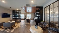 Located at 14750 S Wallin Dr in Plainfield, IL, this single-floor amenity space offers a sophisticated and well-appointed communal environment designed for both relaxation and social gatherings. The interior is immediately distinguished by its eye-catching hexagonal tile flooring, which transitions seamlessly across the open-plan layout, blending earthy tones with lighter accents to create visual depth and texture throughout.

The main lounge area is anchored by a geometric wood coffee table and patterned accent chairs, complemented by a mounted flat-screen TV and modern pendant lighting. Dark, floor-to-ceiling open shelving units serve as stylish dividers, organizing the space into distinct functional zones while maintaining an airy, connected feel. Large windows allow ample natural light to fill the interior throughout the day.

A central dining and work bar stretches through the heart of the space, topped with a crisp white surface and surrounded by sleek black bar-height chairs, making it ideal for casual dining, collaboration, or remote work. Pendant globe lights hang above, adding an upscale touch.

The kitchen area features rich dark cabinetry, open display shelving with gold accents, a mounted TV, and a wood-paneled ceiling that adds warmth. A private meeting room enclosed by black-framed glass doors provides a more intimate gathering option. The overall ambiance is modern, curated, and inviting.