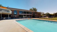 This single-story recreational facility is housed in a classic brick building with a distinctive arched window and a broad span of floor-to-ceiling glass along its front facade, allowing generous natural light to fill the interior and providing clear views of the outdoor pool area. The exterior is framed by mature trees that lend a welcoming, park-like atmosphere to the grounds.

The centerpiece of the outdoor space is a large, well-maintained swimming pool with a clean concrete surround and painted lane markings on the pool floor, complete with entry handrails and safety equipment. Ample deck space surrounds the pool, offering room for seating and recreation under open skies.

Inside, the restroom facilities are thoughtfully designed with functionality and cleanliness in mind. Stainless steel sinks with chrome faucets and crystal-style knobs are mounted above wood-toned cabinetry with warm, honey-oak finishes. Under-sink storage compartments provide practical organization. The restroom stalls are fitted with modern hardware and the overall finish palette — neutral walls paired with wood-grain accents — creates a clean, utilitarian aesthetic that is easy to maintain. Together, the indoor and outdoor components of this facility present a complete, amenity-rich space well-suited for community recreation and aquatic programming.