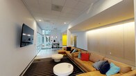 This single-floor communal space is thoughtfully designed to encourage gathering and relaxation, blending contemporary aesthetics with functional amenities. The layout flows naturally from an open lounge area into a bright, glass-fronted zone and a well-appointed kitchenette.

The lounge area anchors the space with a generous caramel-toned sectional sofa dressed in a mix of colorful throw pillows, arranged atop a bold black-and-white striped area rug. Round, sculptural white coffee tables provide a clean contrast. Warm linear lighting along a wood-paneled accent wall adds a refined, welcoming atmosphere, while a wall-mounted flat-screen television makes this an ideal spot for both casual socializing and entertainment.

A floor-to-ceiling glass curtain wall floods the adjacent seating nook with natural light and frames views of the surrounding exterior courtyard. Polished concrete floors and a pendant light above a sleek bistro table with wire-frame chairs lend the space an airy, urban feel.

The kitchenette features a full run of white cabinetry beneath an eye-catching exposed brick accent wall, complemented by a countertop workspace, sink, coffee station, and under-counter beverage refrigerator. A roll-up glass garage door beside the kitchenette opens to the outdoors, enhancing versatility. Bold yellow accent doors and white walls throughout tie the space together with a vibrant, modern energy.