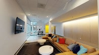 This single-floor communal space is thoughtfully designed to encourage gathering and relaxation, blending contemporary aesthetics with functional amenities. The layout flows naturally from an open lounge area into a bright, glass-fronted zone and a well-appointed kitchenette.

The lounge area anchors the space with a generous caramel-toned sectional sofa dressed in a mix of colorful throw pillows, arranged atop a bold black-and-white striped area rug. Round, sculptural white coffee tables provide a clean contrast. Warm linear lighting along a wood-paneled accent wall adds a refined, welcoming atmosphere, while a wall-mounted flat-screen television makes this an ideal spot for both casual socializing and entertainment.

A floor-to-ceiling glass curtain wall floods the adjacent seating nook with natural light and frames views of the surrounding exterior courtyard. Polished concrete floors and a pendant light above a sleek bistro table with wire-frame chairs lend the space an airy, urban feel.

The kitchenette features a full run of white cabinetry beneath an eye-catching exposed brick accent wall, complemented by a countertop workspace, sink, coffee station, and under-counter beverage refrigerator. A roll-up glass garage door beside the kitchenette opens to the outdoors, enhancing versatility. Bold yellow accent doors and white walls throughout tie the space together with a vibrant, modern energy.