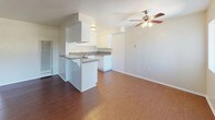 This single-floor space is designed with a practical, open layout that connects the living and kitchen areas seamlessly. The living room features warm wood-look flooring, a ceiling fan with integrated lighting, and large windows fitted with vertical blinds that allow ample natural light to fill the room. A wall-mounted air conditioning unit provides climate control.

The kitchen is positioned adjacent to the living area and is defined by a peninsula counter with granite-style countertops, offering additional workspace and a casual seating option. White cabinetry lines the walls, providing generous storage, while a gas range, overhead ventilation hood, and a double-basin stainless steel sink complete the functional cooking space. The wood-look flooring continues throughout this area.

The bedroom is carpeted and well-lit by natural light streaming through vertical blinds. A ceiling fan and a mirrored sliding closet door add both comfort and a sense of spaciousness to the room.

The bathroom is cleanly finished with white tile surrounds, a shower-over-tub combination, a toilet, and a granite-topped vanity. A frosted privacy window above the tub allows natural light while maintaining privacy. Wood-look flooring ties the bathroom's aesthetic to the rest of the unit.
