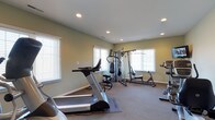 This dedicated fitness room offers a practical and comfortable environment for exercise. The space is finished with soft sage-green walls and neutral carpeted flooring, creating a calm, focused atmosphere suited to physical activity. Recessed ceiling lights provide consistent, even illumination throughout the room, complementing the natural light that filters in through multiple windows dressed with white horizontal blinds. The windows are positioned on two walls, ensuring the space feels open and well-lit without harsh glare. The room accommodates a solid selection of fitness equipment, including at least two treadmills for cardiovascular training and a multi-station cable and weight machine for comprehensive strength training. Additional equipment, such as a rowing or seated exercise machine, is visible along the perimeter. The layout allows for reasonable circulation between machines. White baseboards provide a clean contrast against the wall color, and the overall finish quality reflects a thoughtfully maintained space. This single-level fitness room is well-suited for a variety of workout routines, making it a functional and self-contained exercise area.