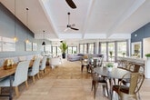 Located at 10235 Boca Entrada Blvd in Boca Raton, Florida, this single-floor community space offers a generously sized, open-plan interior designed for gathering, relaxation, and socializing. The layout is defined by its soaring tray and vaulted ceilings fitted with modern ceiling fans, creating an airy and expansive atmosphere throughout.

Large black-framed glass doors and floor-to-ceiling windows line the perimeter, flooding the interior with natural light and offering views of the surrounding lush, tree-lined landscape. The flooring features a combination of large-format light-toned tile with a decorative parquet-inspired inlay, flowing seamlessly into wood-look planks in the lounge areas.

The space is thoughtfully divided into distinct functional zones. A casual dining area features a long wooden communal table with upholstered chairs and a cushioned banquette, complemented by pendant lighting and coastal-themed wall art. Smaller round tables with metal chairs provide additional seating options. A comfortable lounge area includes a sectional sofa and accent seating arranged around a wall-mounted television set against a textured blue accent wall.

A built-in workstation area with desktop computers offers a dedicated workspace, while a service kitchen with stainless steel appliances and white cabinetry is accessible nearby. Lush tropical potted plants add vibrancy and a Florida-inspired ambiance throughout the space.