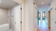 This contemporary single-floor studio is thoughtfully designed to maximize every inch of its compact footprint. The open-plan kitchen and living area serves as the heart of the space, featuring warm natural hardwood flooring that runs throughout and large black-framed floor-to-ceiling windows that flood the interior with abundant natural light and offer views of the surrounding urban streetscape. The kitchen is outfitted with sleek, flat-front white cabinetry extending from floor to ceiling, a marble-style backsplash, stainless steel Bosch appliances including a gas cooktop and double wall oven, and an undermount sink with brushed nickel fixtures — providing a clean, modern aesthetic. The adjacent living area is bright and open, with recessed lighting complementing the natural light. A hallway connects the main living space to the private areas and features mirrored sliding doors, adding depth and dimension. The full bathroom is elegantly finished with large-format neutral tiles, a soaking tub with overhead shower, a floating white vanity with an integrated sink, and a backlit mirror, creating a serene and spa-like atmosphere. The consistent white palette and modern finishes throughout give the space a cohesive, refined character.