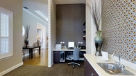 This single-floor space combines functional design with refined aesthetics throughout its layout. A standout feature is the dedicated built-in workstation nook, set against a striking floor-to-ceiling textured accent wall in deep charcoal tones. The workspace is equipped with built-in shelving, dark cabinetry with ample storage, and a clean countertop surface that accommodates a full desktop setup — making it ideal for productivity within a polished environment. The area transitions seamlessly into a wet bar or kitchenette zone, featuring dark lower cabinetry, a light countertop, and a sink, adding a layer of everyday convenience. Decorative elements, including an ornate vase with sculptural branches and a mosaic-patterned accent panel, lend the space a sophisticated, curated feel. The flooring shifts from warm hardwood to a natural fiber area rug, delineating the workstation zone while maintaining visual cohesion. A structural column with classic white detailing adds architectural character and subtly separates the work area from an adjoining room visible in the background, which appears to host a dining or sitting area bathed in warm ambient light. The overall ambiance is professional yet inviting, balancing bold design choices with functional, well-organized interiors.