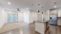 This single-floor residence offers a well-designed layout encompassing two bedrooms, two full bathrooms, and two closets within a thoughtfully utilized footprint. The open-concept kitchen and living area serves as the centerpiece of the home, featuring wood-look plank flooring that extends throughout the main living spaces. The kitchen is appointed with two-tone cabinetry — white upper cabinets paired with wood-grain lower cabinets — complemented by a herringbone tile backsplash, granite countertops, stainless steel appliances, and a large island with a dark granite surface, undermount sink, and pendant lighting above. The living area transitions seamlessly from the kitchen, featuring a ceiling fan, recessed lighting, and a glass door providing natural light and outdoor access. Both bedrooms are finished with plush carpet flooring; the primary bedroom includes a ceiling fan and exterior glass door access. Each bathroom features a wood-grain vanity with granite countertops, an illuminated mirror, matte black fixtures, and a tub-shower combination with tile surround. The unit maintains a clean, contemporary aesthetic throughout with crisp white walls and consistent modern detailing.