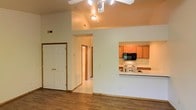 This single-story space showcases an open, functional layout designed for comfortable everyday living. The living area features warm hardwood-style flooring and a vaulted ceiling equipped with a ceiling fan and track lighting, adding both airflow and brightness to the space. A pass-through counter connects the living area to the kitchen, creating an open, communicative flow between the two spaces. The kitchen is outfitted with honey-toned wood cabinetry, a white electric range, a black microwave, and a double sink with light-colored countertops, offering a practical and well-organized cooking environment. Light tile flooring in the kitchen and hallway provides a clean contrast to the darker floors in the living area. A hallway visible from the main living space leads to additional rooms, suggesting a thoughtful floor plan that separates the living and private areas. The entryway, framed by a classic six-panel white door, provides a welcoming transition into the home. Neutral wall tones throughout contribute to a bright, airy ambiance that complements the warm wood accents found in both the flooring and cabinetry.