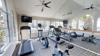 This fitness center is designed to support a well-rounded cardio workout routine within a bright, welcoming environment. The space is outfitted with a comprehensive selection of cardio equipment, including treadmills, elliptical trainers, and stationary bikes, arranged to allow comfortable use and movement between machines. Patterned low-pile carpet in neutral tones covers the floor, providing both durability and a polished aesthetic. A full-length wall mirror with a gold-tone rail runs along one side of the room, visually expanding the space and allowing users to monitor their form during exercise. Large windows along the perimeter flood the interior with natural light and offer views of the surrounding exterior, contributing to an open and airy atmosphere. Ceiling fans are mounted overhead to maintain comfortable airflow throughout workouts. Wall-mounted flat-screen televisions are positioned for easy viewing from the equipment. The textured ceiling adds a classic finishing touch, while the overall layout and design of the space reflect a clean, functional approach to community fitness amenities.