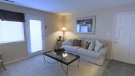 This single-floor space centers around a welcoming living room designed with a neutral, understated palette. The room is finished with wall-to-wall carpeting in a soft beige tone, contributing to a warm and comfortable atmosphere. Walls are painted in a light, warm neutral, creating a cohesive and calming environment throughout.

A large, cushioned sofa in off-white serves as the focal point of the living area, dressed with decorative brown and cream accent pillows. A glass-top coffee table with an ornate wrought-iron base sits at the center, adding a touch of elegance to the space. Ambient lighting is provided by a classic table lamp positioned on a side console, casting a soft, inviting glow across the room.

A framed piece of wall art adds visual interest above the sofa, while a full-length door with sidelights along one wall allows natural light to filter into the space, brightening the interior. The overall design balances simplicity with comfort, making the living area feel both functional and inviting for everyday use.