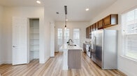 This well-designed single-floor space offers a clean, contemporary aesthetic throughout, unified by light wood-tone luxury vinyl plank flooring and crisp white walls that create a bright, airy atmosphere. The open-concept kitchen serves as the heart of the home, anchored by a generous center island with a white quartz countertop, an undermount sink, and a stylish pendant light overhead. Rich walnut-toned cabinetry lines the kitchen walls, complemented by a full suite of stainless steel appliances including a French door refrigerator, electric range with hood, and built-in microwave. A white subway tile backsplash adds a classic touch, while shuttered windows allow for natural light and privacy. Adjacent to the kitchen, a large open pantry with built-in shelving provides excellent storage and organization. The living area is open and versatile, bathed in natural light from multiple windows. A dedicated bedroom features a window with shutters and direct access to a spacious walk-in closet, outfitted with wrap-around shelving, hanging rods, and built-in vertical storage units. The bathroom is finished with warm wood cabinetry, quartz countertops, a wall-mounted mirror, and a walk-in shower enclosed by sleek black-framed glass doors with white tile surround.