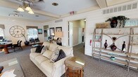 This single-floor space offers a warm and inviting atmosphere enhanced by thoughtful design details throughout. The interior is characterized by white shiplap-style wall paneling and beamed ceilings that lend a classic, cottage-inspired aesthetic across the entire layout.

The centerpiece of the main living area is a commanding floor-to-ceiling stone fireplace, featuring a broad slate hearth and a white wood mantle accented by wall sconces and framed artwork above. Comfortable seating, including a plush neutral sofa with decorative pillows and a wingback armchair, is arranged to take full advantage of this focal point. A glass-topped coffee table anchors the seating arrangement, while an open metal shelving unit displays decorative accessories throughout the reception area.

Adjacent to the main lounge, a functional workspace is equipped with a desk, office seating, and a large decorative wall clock, creating a productive yet aesthetically cohesive area. Ceiling fans provide comfortable airflow throughout.

A dining area features a round glass-top table surrounded by dark wood chairs, positioned beside a sliding glass door that floods the space with natural light and provides direct access to the exterior. Patterned carpeting runs throughout, tying the spaces together cohesively. Additional rooms are accessible via interior doorways, rounding out this well-appointed and versatile space.